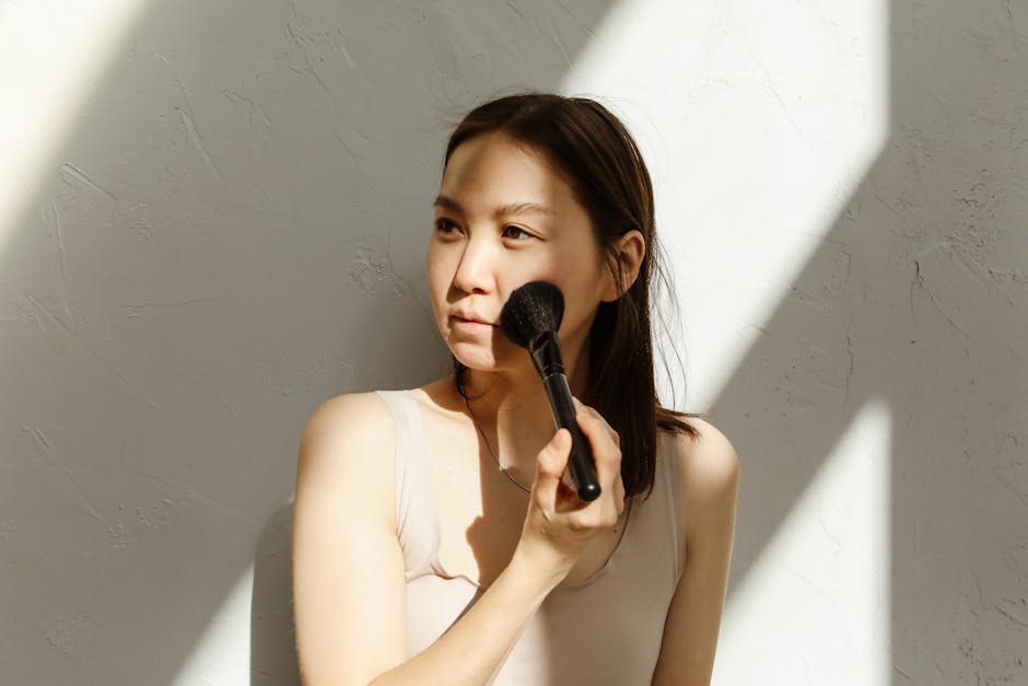A young Asian woman applying makeup with a brush in natural lighting.